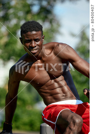 Muscular young man exercising at the sports field. African man looking to the side while doing horizontal bar exercises. Shirtless male model exercising outdoors. 80400407