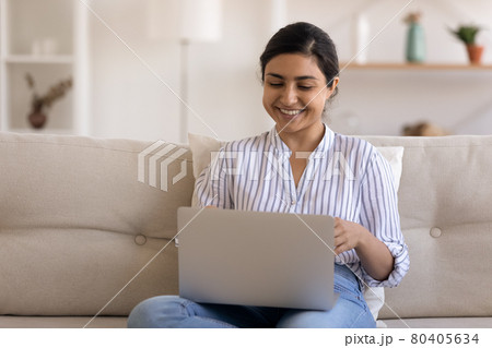 Smiling Indian woman using laptop, having fun, sitting on couch 80405634