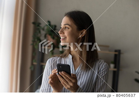 Dreamy beautiful woman holding smartphone standing indoor looks out window Dreamy beautiful woman holding smartphone standing indoor looks out window 80405637