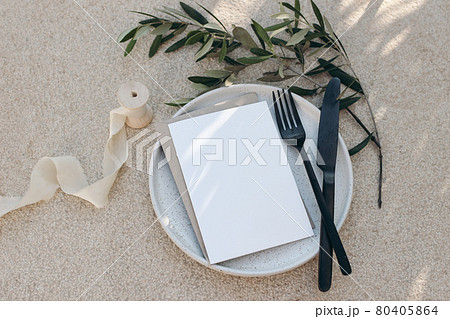 Festive wedding summer table setting. Black cutlery, olive branch, silk ribbon on dotted white dinner plate. Beige concrete background. Blank greeting card, invittation mockup. Flat lay, top view. Festive wedding summer table setting. Black cutlery, olive branch, silk ribbon on dotted white dinner plate. Beige concrete background. Blank greeting card, invittation mockup. Flat lay, top view. 80405864