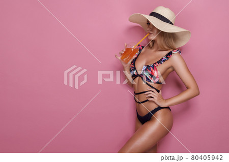 stylish woman in straw hat, sunglasses and swimsuit holding glass with orange juice on pink background. 80405942
