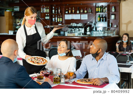 Polite waitress in protective mask bringing ordered pizza to friends visited restaurant Polite waitress in protective mask bringing ordered pizza to friends visited restaurant 80409992