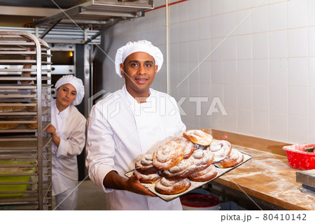 Smiling bakery worker carrying tray with baked ensaimadas Smiling bakery worker carrying tray with baked ensaimadas 80410142