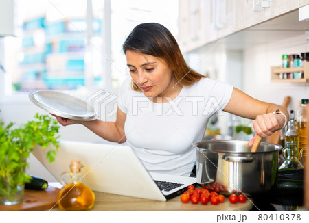 Woman cooking soup and watching movie at laptop 80410378