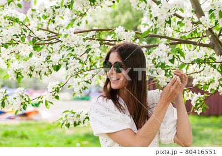 Attractive and beautiful young woman in sunglasses. Young woman caucasian ethnicity. Smiling and happy female is posing in white clothes. Cheerful pretty woman on blooming apple tree background. Attractive and beautiful young woman in sunglasses. Young woman caucasian ethnicity. Smiling and happy female is posing in white clothes. Cheerful pretty woman on blooming apple tree background. 80416551