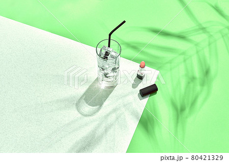 High angle view of lipstick and drinking glass with ice on table. Green and white background with shadow from a palm leaf 80421329