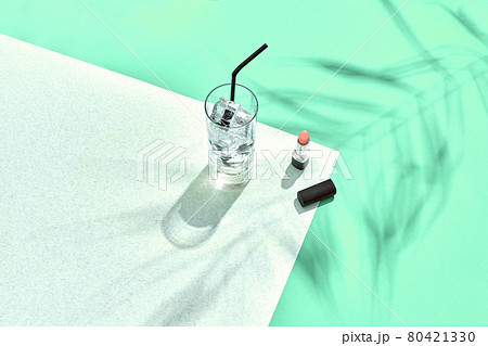 High angle view of lipstick and drinking glass with ice on table. Blue and white background with shadow from a palm leaf 80421330