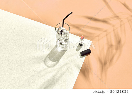 High angle view of lipstick and drinking glass with ice on table. Beige and white background with shadow from a palm leaf 80421332