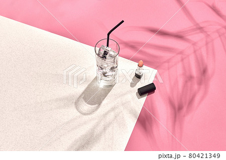 High angle view of lipstick and drinking glass with ice on table. Pink and white background with shadow from a palm leaf 80421349