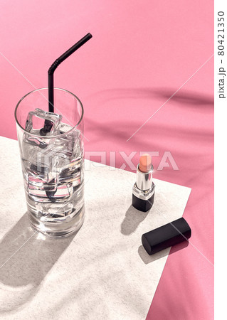 High angle view of lipstick and drinking glass with ice on table. Pink and white background with shadow from a palm leaf 80421350