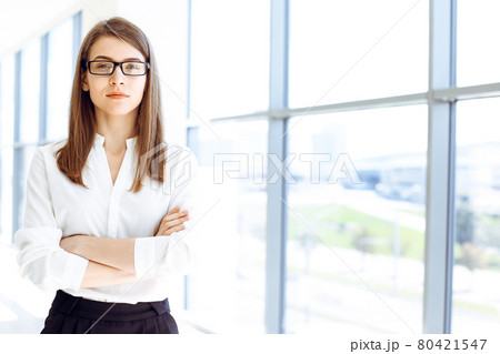 Beautiful female specialist with arms crossed standing in modern office and smiling charmingly. Working on design, data analysis, plan strategy. Business people concept Beautiful female specialist with arms crossed standing in modern office and smiling charmingly. Working on design, data analysis, plan strategy. Business people concept 80421547