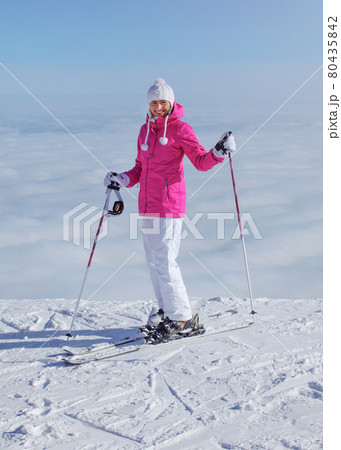 Woman in pink jacket, ski poles and skis, standing at the edge of hill, only clouds below, looking back, smiling Woman in pink jacket, ski poles and skis, standing at the edge of hill, only clouds below, looking back, smiling 80435842