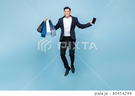 Excited young handsome Asian man holding mobile phone and shopping bags jumping Excited young handsome Asian man holding mobile phone and shopping bags jumping 80454299