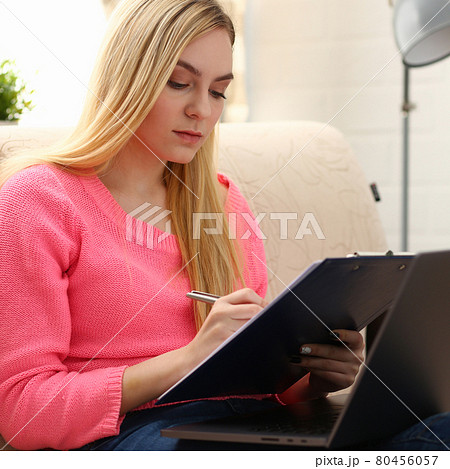 young beautiful blond woman sit on the sofa in livingroom hold binder in arms work on laptop 80456057