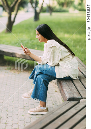 A girl writes SMS messages or uses applications on a smartphone while sitting on a bench in the park A girl writes SMS messages or uses applications on a smartphone while sitting on a bench in the park 80458687