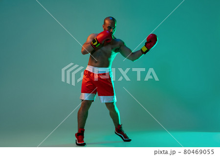 Portrait of one professional boxer in red gloves training, exercising over blue green background in neon light. Full length 80469055