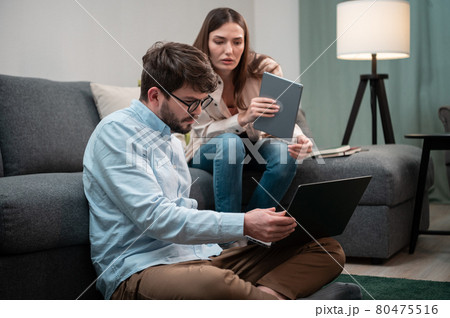 A man and a woman from a remote home office are having intense phone conversations from the home office, sitting on and next to the couch during lockdown A man and a woman from a remote home office are having intense phone conversations from the home office, sitting on and next to the couch during lockdown 80475516