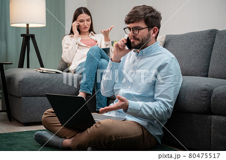 A man and a woman from a remote home office are having intense phone conversations from the home office, sitting on and next to the couch during lockdown A man and a woman from a remote home office are having intense phone conversations from the home office, sitting on and next to the couch during lockdown 80475517