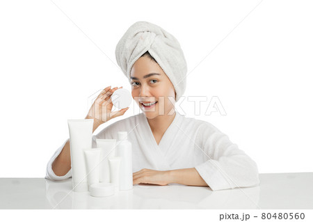 Smiling beautiful girl model with towel on head holding cream pot bottle with various skin care products on table Smiling beautiful girl model with towel on head holding cream pot bottle with various skin care products on table 80480560