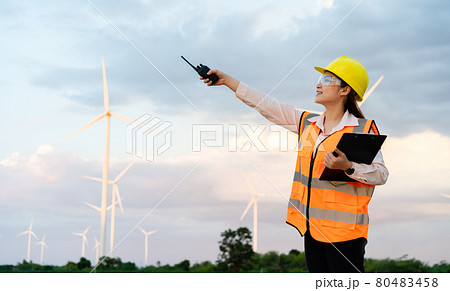 female engineer using walkie talkie and reading clipboard to checking system against wind turbine farm 80483458