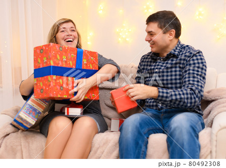 portrait of adult romantic couple, sitting with gifts on a couch at home, a woman takes away all the gifts from a man, the photo is like a joke, holiday lights on a wall 80492308