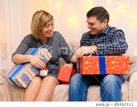 portrait of adult romantic couple, sitting with gifts on a couch at home, holiday lights on a wall 80492311