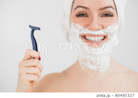 Cheerful caucasian woman with a towel on her head and shaving foam on her face holds a razor on a white background 80495327
