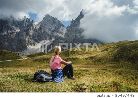 Dolomites. Woman sitting on the grass enjoying Baita Segantini mountain with Cimon della Pala peak, refuge and lake in background. Rolle pass, Trentino province, Italy, Europe Dolomites. Woman sitting on the grass enjoying Baita Segantini mountain with Cimon della Pala peak, refuge and lake in background. Rolle pass, Trentino province, Italy, Europe 80497448