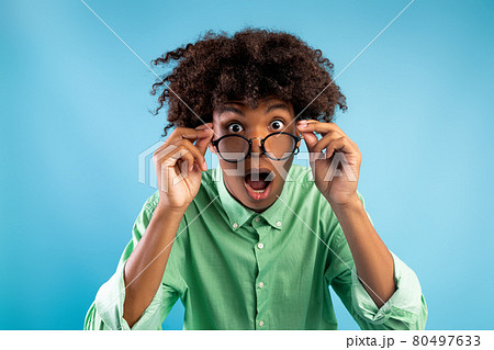 Emotional african american teen guy in glasses opening mouth in surprise, looking at camera over blue background 80497633