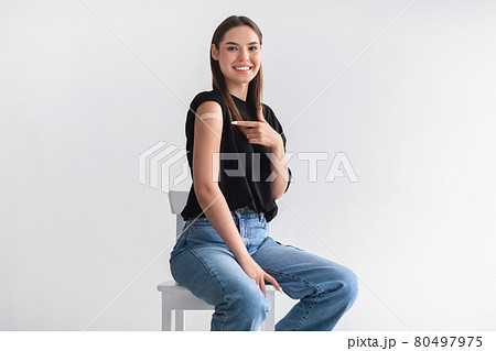 Happy young female patient pointing at her arm with band aid after coronavirus vaccine shot on light studio background 80497975