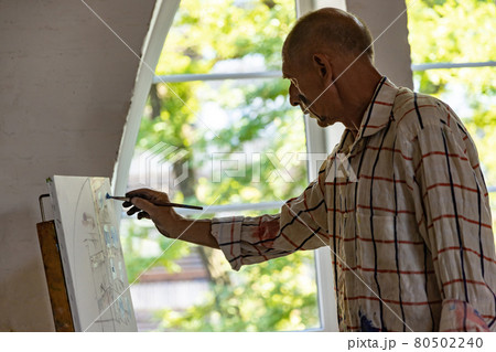 Mature or senior grey painter with a brush standing in front of camera. Serious man in a paint-stained shirt working at studio or home 80502240