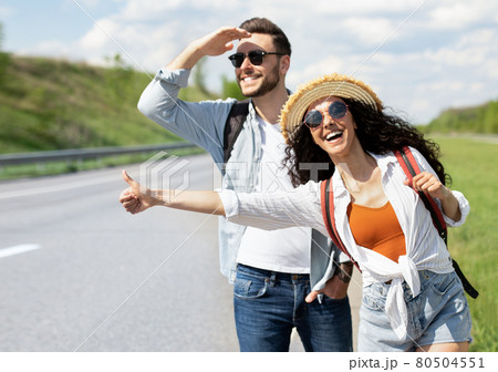Happy young coupe flagging down car, needing ride, hitchhiking on roadside outdoors Happy young coupe flagging down car, needing ride, hitchhiking on roadside outdoors 80504551