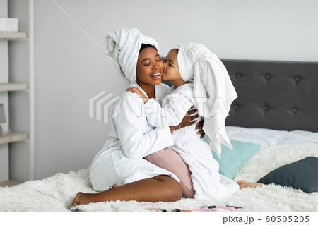 Full length portrait of little girl kissing her black mother on makeup day, wearing bath robes and towels in bedroom 80505205