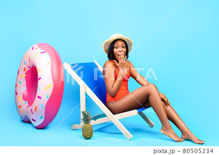 Oops. Shy black lady in bikini chilling in lounge chair, covering her mouth, making mistake on blue studio background Oops. Shy black lady in bikini chilling in lounge chair, covering her mouth, making mistake on blue studio background 80505234