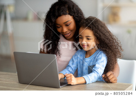 Home schooling during covid concept. Cheerful black mom helping her daughter with online homework on laptop pc Home schooling during covid concept. Cheerful black mom helping her daughter with online homework on laptop pc 80505284