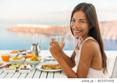 Breakfast woman eating brunch at luxury travel hotel restaurant drinking coffee cup over Mediterranean view from outside balcony. Greek healthy food Santonini vacation. Breakfast woman eating brunch at luxury travel hotel restaurant drinking coffee cup over Mediterranean view from outside balcony. Greek healthy food Santonini vacation. 80505408