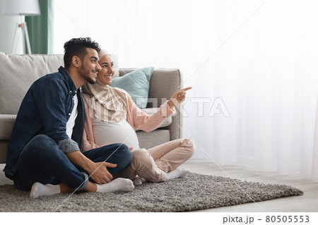 Portrait Of Happy Pregnant Middle-Eastern Couple Resting On Floor In Living Room 80505553