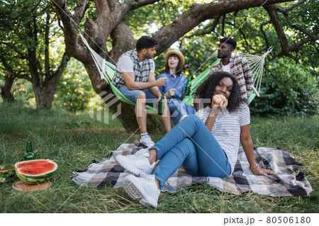 Positive black woman eating croissant on picnic with friends 80506180