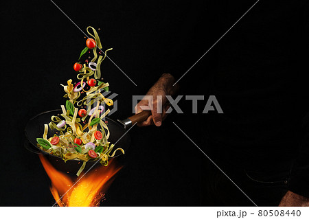 Unrecognizable man holding wok pan above fire and cooking pasta with cherry tomatoes, onion and basil against black background. Close up Unrecognizable man holding wok pan above fire and cooking pasta with cherry tomatoes, onion and basil against black background. Close up 80508440