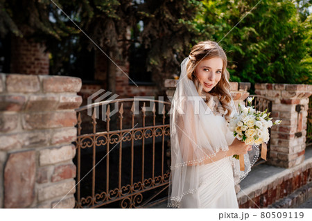 gentle and beautiful bride in a veil with a bouquet of flowers at the fence. gentle and beautiful bride in a veil with a bouquet of flowers at the fence. 80509119
