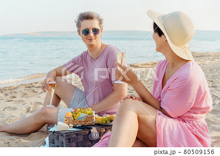 Laughing couple having a picnic at the seaside beach. Man and woman in love holding glasses with wine or champagne, celebrating the moment with sea background. Romantic date, enjoy vacation concept. 80509156