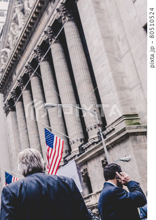Businessman talking on the phone on Wall street in New York with American flags and New York Stock Exchange in background. 80510524
