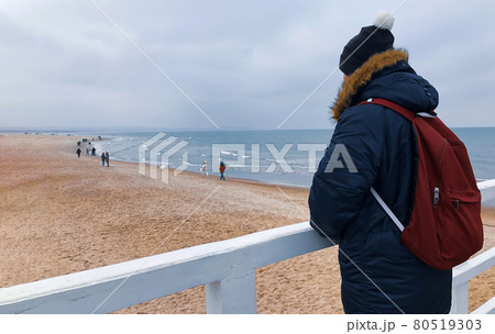 A young man in winter in a jacket, a hat and a backpack stands on the pier and looks at the Baltic Sea 80519303
