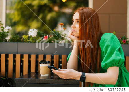 Side view of sad redhead young woman holding mobile phone, sitting at table with coffee cup in outdoor cafe terrace in sunny summer day 80522243