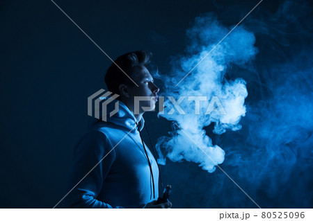 A young guy smokes a vape in the studio in neon light. 80525096