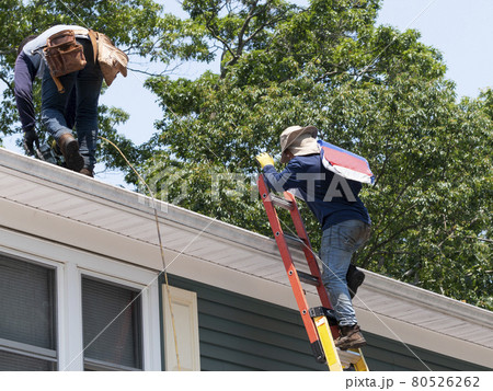 contractor climbing ladder with roof shingles while another nail the shingles on 80526262