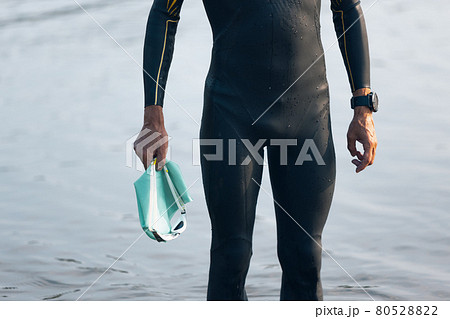 Professional triathlete before swimming in river's open water. Man wearing swim equipment practicing triathlon on the beach in summer's day. 80528822