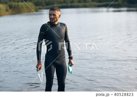 Professional triathlete before swimming in river's open water. Man wearing swim equipment practicing triathlon on the beach in summer's day. Professional triathlete before swimming in river's open water. Man wearing swim equipment practicing triathlon on the beach in summer's day. 80528823