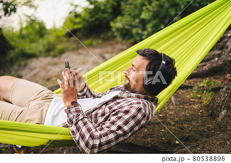 Young caucasian man resting in hammock, listening to music on headphones and using smartphone afterwards on bicycle in forest near the lake. Audio healing. Idyllic place. Travel, camping in nature Young caucasian man resting in hammock, listening to music on headphones and using smartphone afterwards on bicycle in forest near the lake. Audio healing. Idyllic place. Travel, camping in nature 80538896