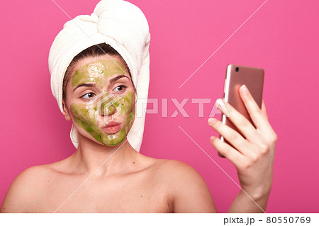 Emotional adorable cosmetologist making selfie for social networking sites, holding smartphone in her hand, head wrapped in towel, posing half naked, having facial green mask applied on skin. Emotional adorable cosmetologist making selfie for social networking sites, holding smartphone in her hand, head wrapped in towel, posing half naked, having facial green mask applied on skin. 80550769